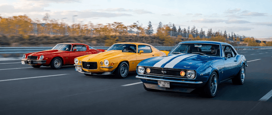 Three classic Chevrolet Camaros—a red, yellow, and blue model—drive side by side on a highway with autumn trees and clear skies in the background.