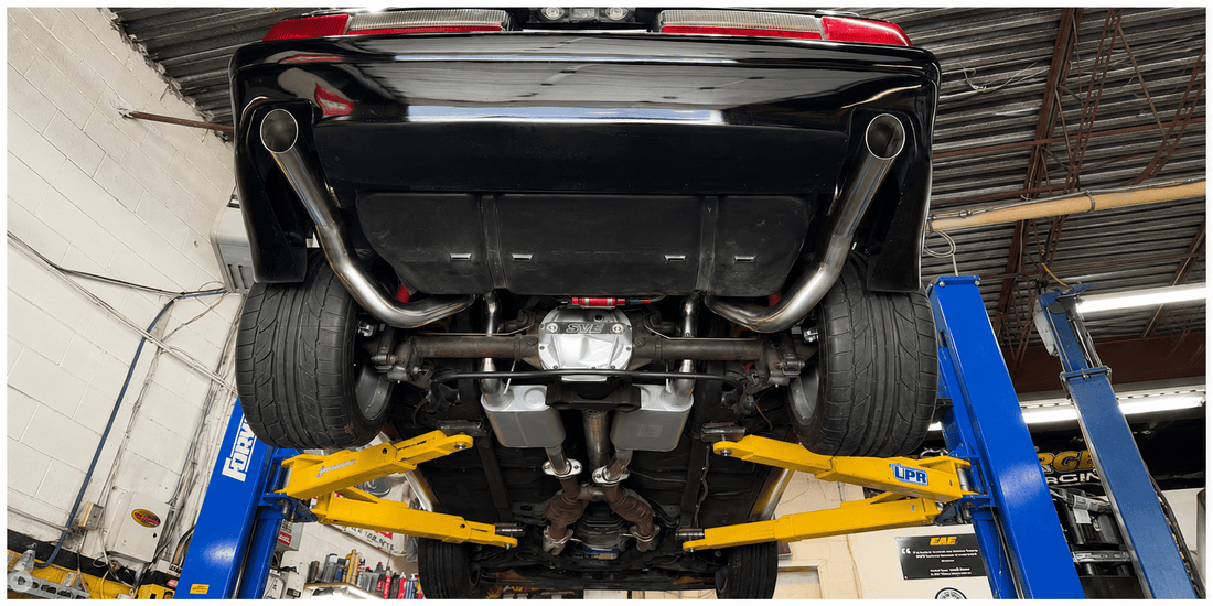 Underside of a lifted black car on a shop lift, showing exhaust, differential, and suspension, in a well‑lit workshop.