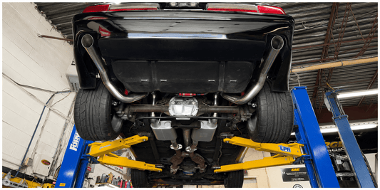 Underside of a lifted black car on a shop lift, showing exhaust, differential, and suspension, in a well‑lit workshop.