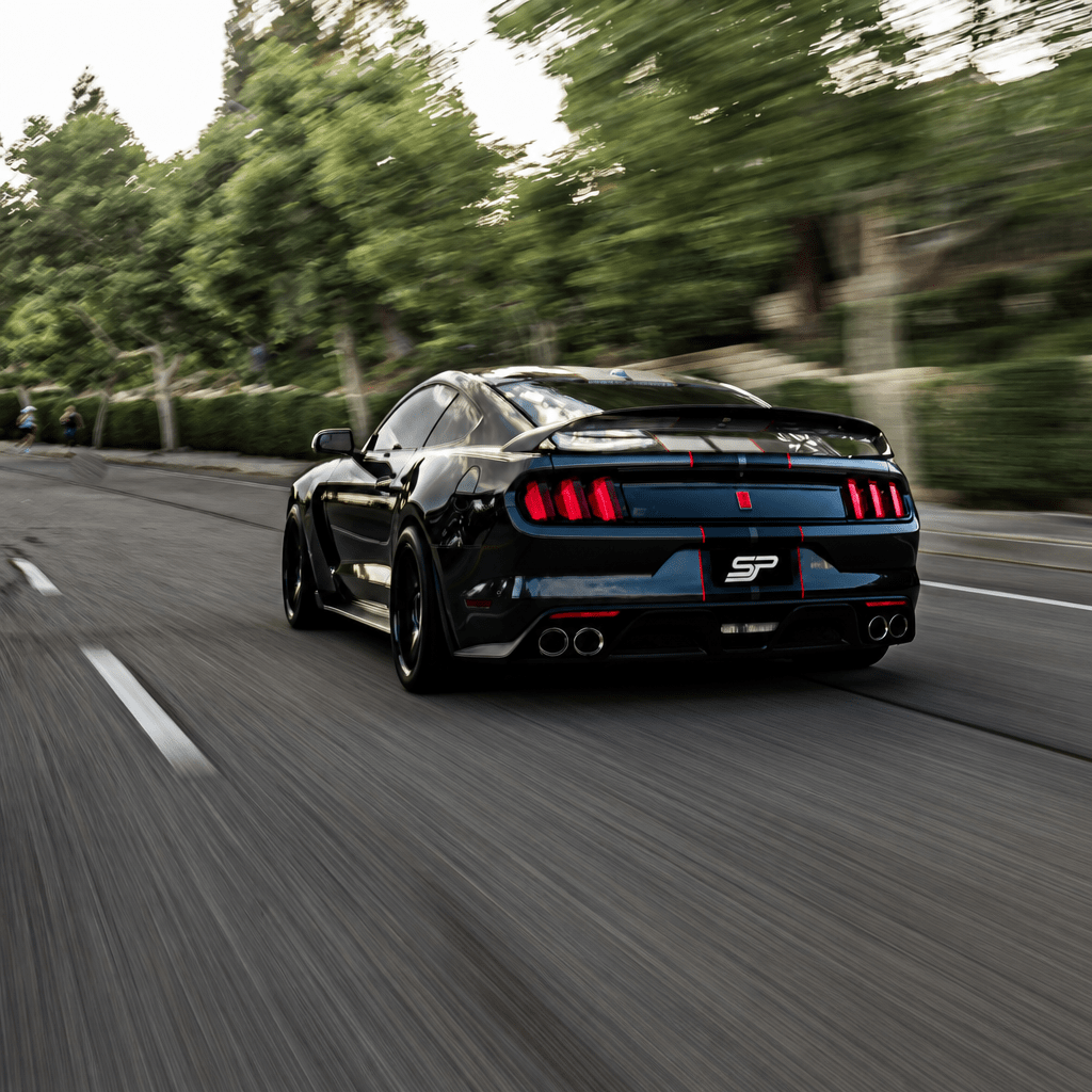Black Ford Mustang driving at speed, captured from behind with taillights, rear spoiler, and quad exhaust on a roadway
