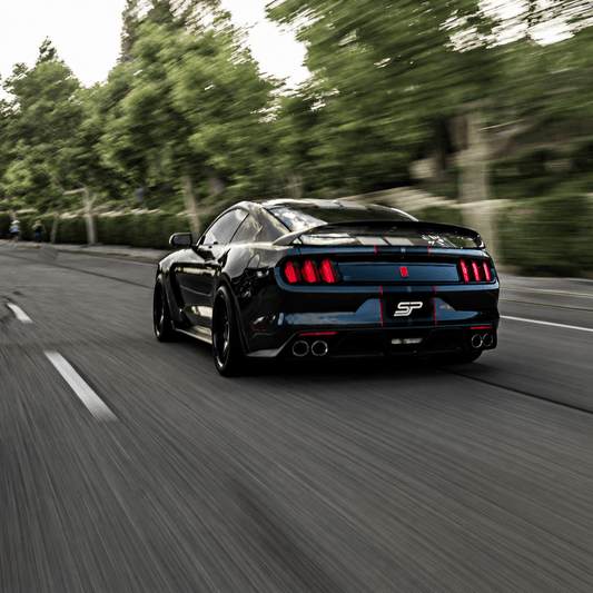 Black Ford Mustang driving at speed, captured from behind with taillights, rear spoiler, and quad exhaust on a roadway