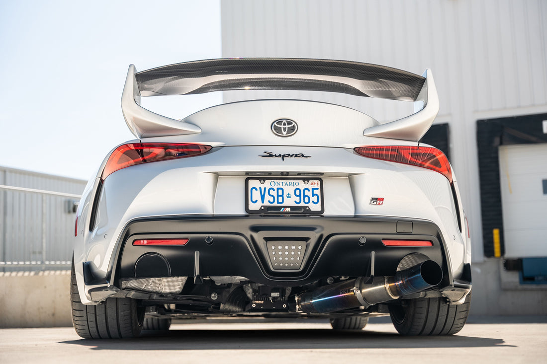 Rear view of a white Toyota Supra with a titanium exhaust, parked outdoors near a building, showcasing its sporty design.