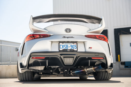 Rear view of a white Toyota Supra with a titanium exhaust, parked outdoors near a building, showcasing its sporty design.