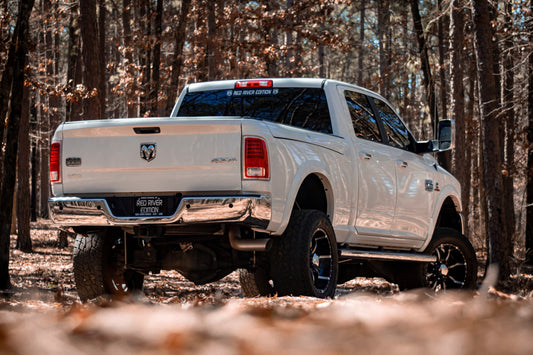 A white lifted Ram 2500 pickup truck with large off‑road tires and black wheels is parked in a forested area.
