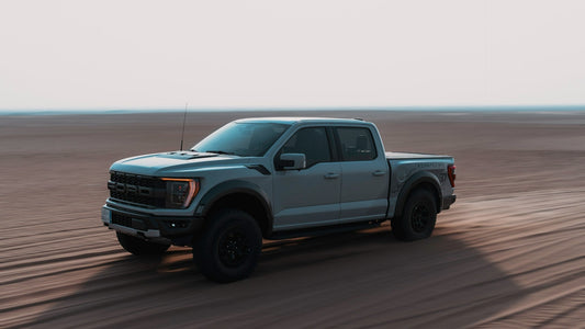 Light gray Ford Raptor driving across open desert terrain, kicking up dust and showcasing its off‑road capability.