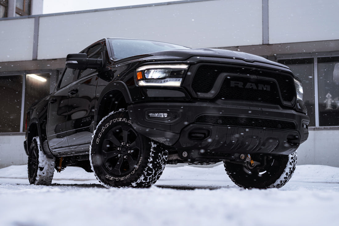 Black RAM pickup parked in falling snow, with rugged tires and a modern building behind it.